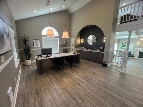 A welcoming leasing office here at Island Park featuring wood-style flooring, a central desk with computer stations and chairs, and modern pendant lighting overhead. The space includes a curved accent wall with a built-in counter, decorative mirror, greenery, and an open layout that connects to the clubhouse seating area beyond.