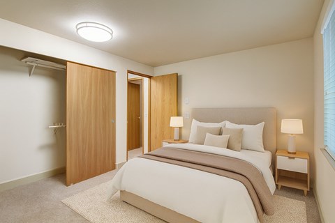 A calm bedroom here at Island Park featuring a bed with neutral bedding and layered pillows, a soft area rug over plush carpeting, and matching bedside tables with modern lamps. The space includes a sliding closet with wood doors, light-colored walls, and a wide window with blinds that brings in natural light.