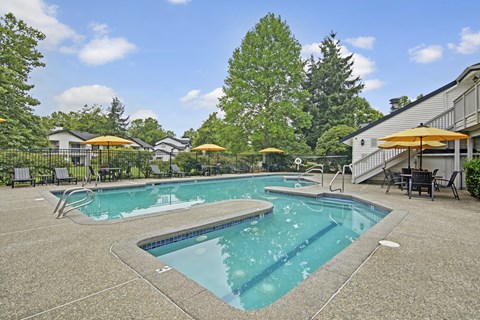 A refreshing outdoor swimming pool here at Island Park featuring a spacious concrete deck, a separate shallow pool area, and multiple lounge chairs with bright umbrellas. The fenced pool area is surrounded by mature trees, landscaped greenery, and nearby community buildings, creating a relaxing setting for residents.