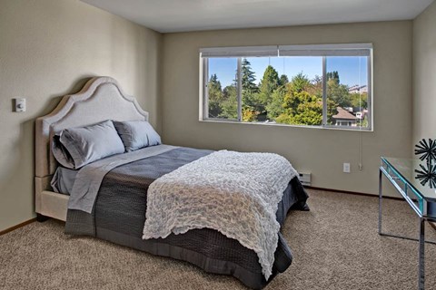 A bright bedroom here at Leilani on Greenwood featuring soft carpeted flooring, neutral walls, and a bed with layered gray and white bedding. A large window with blinds fills the room with natural light and offers leafy neighborhood views, while the open layout provides comfortable space for bedroom furnishings.