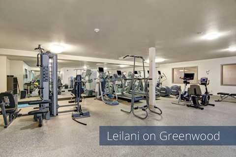 A spacious fitness center here at Leilani on Greenwood featuring a variety of cardio machines, strength training equipment, and free weights arranged throughout an open, well-lit room with neutral flooring and ceiling lights, creating a convenient on-site workout space for residents.