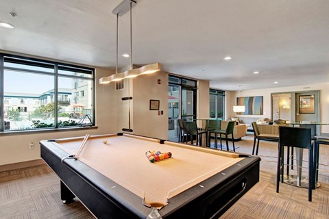A modern resident lounge here at Leilani on Greenwood featuring a billiards table centered beneath contemporary pendant lighting, with large windows providing natural light, adjacent seating and dining areas, and a comfortable open layout designed for socializing and relaxation.