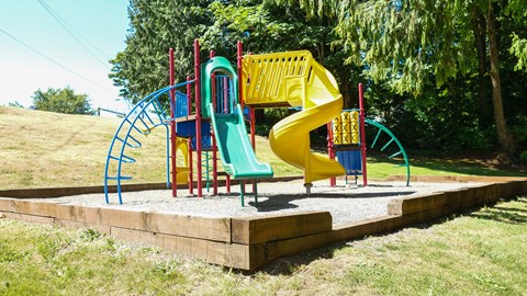 A colorful outdoor playground here at Regency Woods featuring a raised play structure with red support posts, a green straight slide, a yellow spiral slide, climbing ladders, and activity panels, all set on a gravel play surface bordered by timber edging and surrounded by open grassy areas and mature evergreen trees.