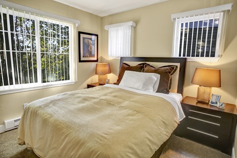 A cozy bedroom here at Regency Woods featuring a comfortable bed with layered neutral bedding, matching nightstands with warm table lamps, soft carpeted flooring, and multiple windows with vertical blinds that allow natural light while offering leafy outdoor views.