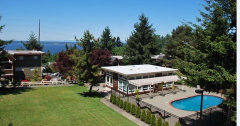 A scenic outdoor pool area here at Soundview with clear blue water, clubhouse, open green lawn, and beautiful water views surrounded by trees.