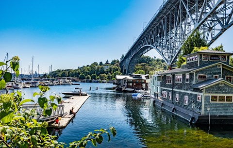 A bridge spans across a calm body of water with houses on the right and boats in the distance.