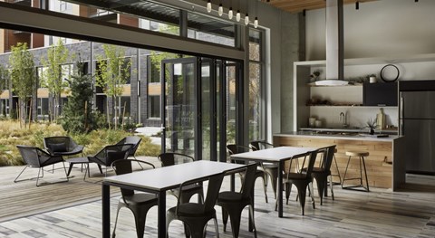 A stylish resident lounge here at Uptown at Kirkland Urban with modern dining tables, a sleek kitchen, and large glass doors opening to a landscaped patio, creating a bright, social space for relaxing or entertaining.