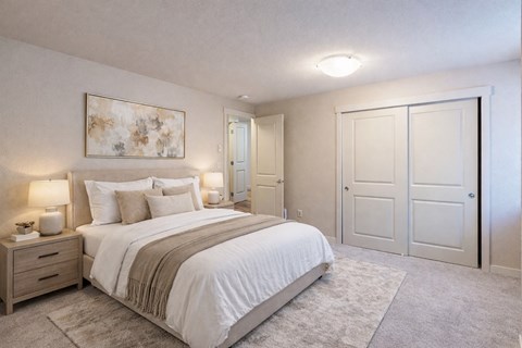 A bedroom here at Vermont featuring soft carpet, large closet doors, neutral bedding, bedside tables with lamps, and abstract wall art, creating a cozy, calm, and inviting retreat.