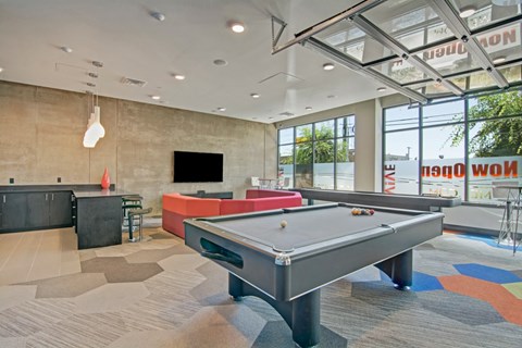 A game lounge here at Vive featuring a sleek pool table, vibrant red sofa, dark entertainment bar, modern pendant lights, and large bright windows creating a stylish, inviting atmosphere.