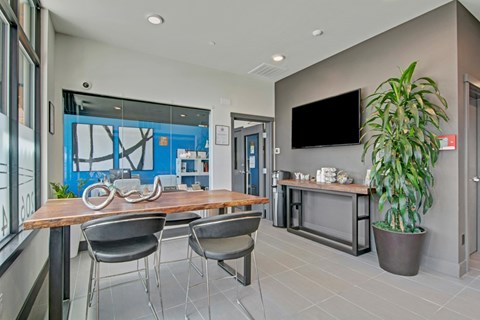 A lounge here at Vive featuring a warm wood table, black chairs, large leafy plant, sleek wall-mounted TV, and modern decor, creating a bright, welcoming space for relaxing or meeting.