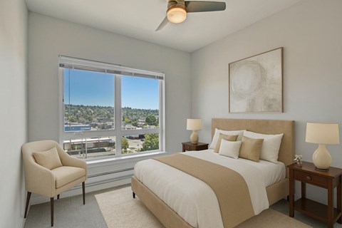 A bright bedroom here at Vive featuring soft neutral bedding, warm wood nightstands, a cozy accent chair, and a large window that fills the inviting space with natural light and scenic neighborhood views.