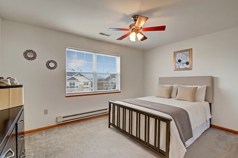 A bright bedroom here at Arbor Ridge featuring white bedding with beige accents, a dark wood bed frame, and a matching dresser. Large window and soft carpet create a calm, inviting retreat.