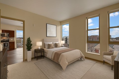 A bright bedroom here at Avenir featuring soft neutral bedding, warm beige walls, and large sunny windows. Matching nightstands with lamps and a cozy accent chair create a calm, welcoming, and comfortable retreat.