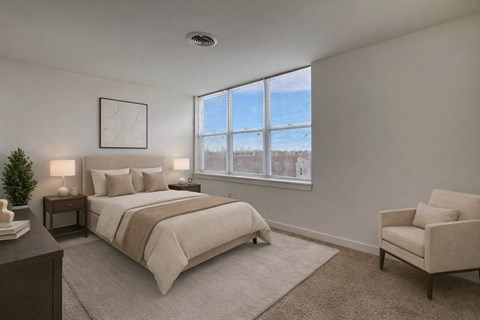 A bright bedroom here at Latitude featuring neutral bedding, dark wood nightstands, and a soft area rug, complemented by a cozy accent chair and large sunlit windows creating a calm, welcoming atmosphere.