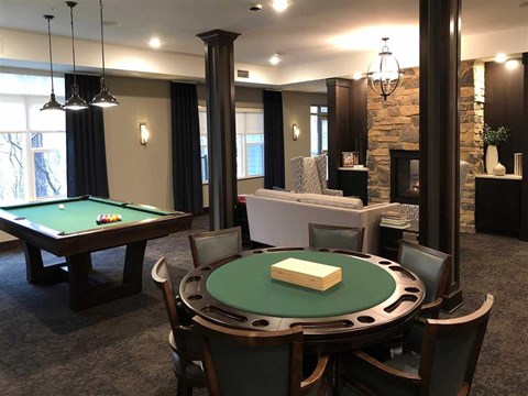 A game room here at Oaks of Shorewood featuring a green poker table, classic pool table, stone fireplace, and comfortable seating, creating a warm, inviting, and social entertainment space.