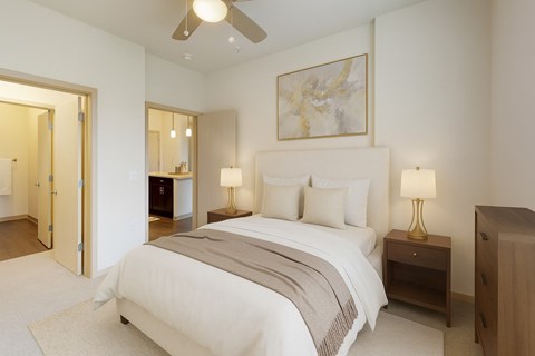 A bright bedroom here at Royster Commons with soft carpet, cream walls, and a ceiling fan for added comfort. A connected bathroom and warm lighting create a comfortable and inviting atmosphere.