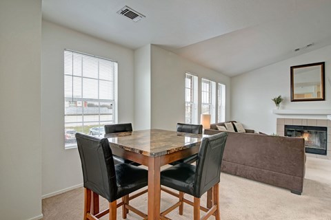A bright dining area here at St. James Place with soft beige carpet, large windows, and a modern dining table. A nearby fireplace and open living space create a warm and inviting atmosphere.
