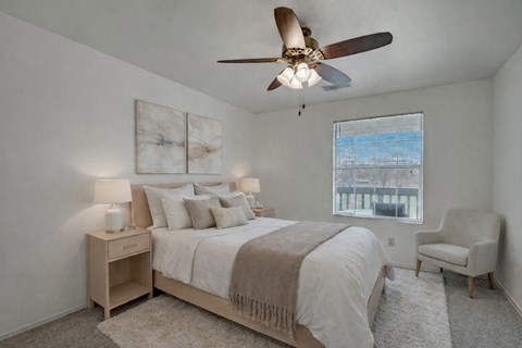 A bright bedroom here at St. James Place with soft beige carpet, cream walls, and a large window that brings in natural light. A ceiling fan and cozy layout create a comfortable and inviting atmosphere.