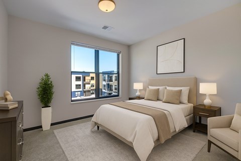 A peaceful bedroom here at The Lafayette featuring soft neutral bedding, matching nightstands with lamps, a light area rug, a cozy accent chair, and a large window bringing in bright, refreshing natural light.