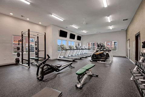 A bright fitness center here at The Park featuring modern equipment, rubber flooring, large windows, multiple treadmills, weight machines, benches, and mounted TVs, creating an energetic and inviting workout space.