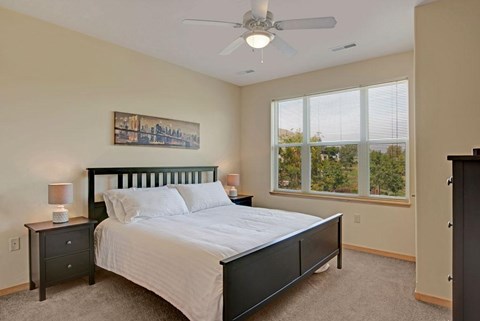 A bedroom here at Van Buren Place featuring a dark wood bed frame, soft carpet flooring, large bright windows with peaceful views, and a clean, cozy layout that feels warm and inviting.