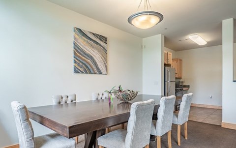 A dining area here at Van Buren Place featuring a large wood table, soft gray upholstered chairs, and a warm overhead light that makes the space feel open, inviting, and perfect for gatherings.