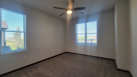 A room with a carpeted floor, a ceiling fan, and two windows.