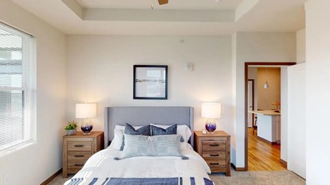 A bedroom here at Van Buren West featuring a soft gray upholstered bed, matching wood nightstands, purple glass lamps, and light neutral bedding for a bright, calm, and inviting space.