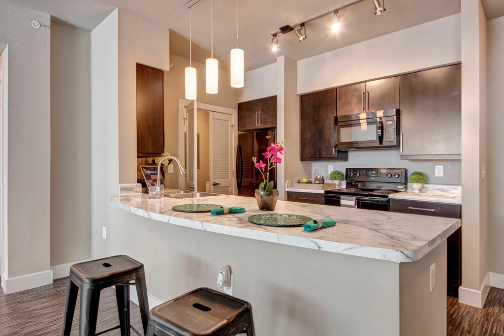 Modern kitchen here at the Enclave at Brookside Apartments with dark wood cabinets and marble countertop. Three pendant lights hang above, with two bar stools beneath. A pink orchid adds color.
