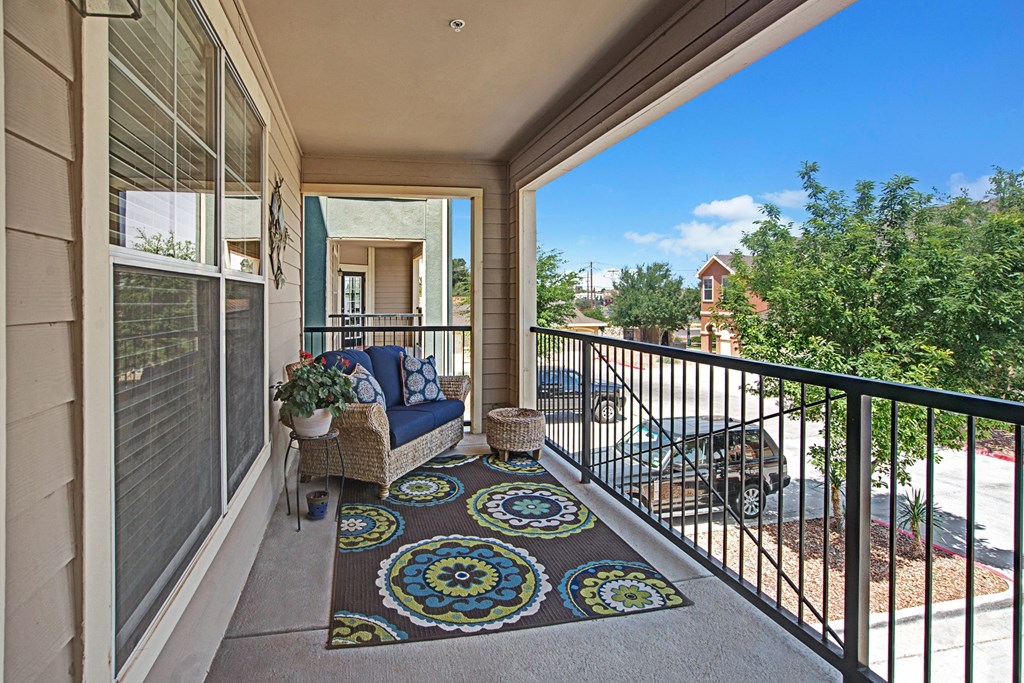 Cozy balcony here at The Palms at Briarwood Apartment Homes with wicker seating, vibrant floral rug, and potted plant. Overlooks a sunny residential neighborhood with trees and parked cars.