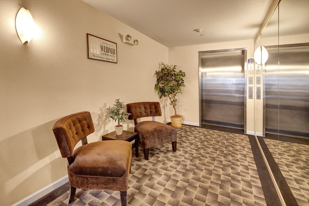 Lobby scene at Admirals  Cove Apartments featuring two brown chairs and potted plants on a patterned carpet near a metallic elevator. Soft lighting creates a warm, welcoming atmosphere.