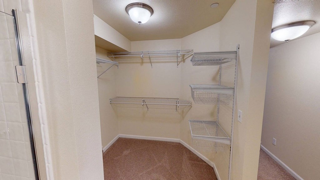 A spacious walk-in closet with beige walls and carpeted floor, featuring metal wire shelving and baskets. Soft, warm lighting from a ceiling fixture. Located at Admirals Cove Apartments.