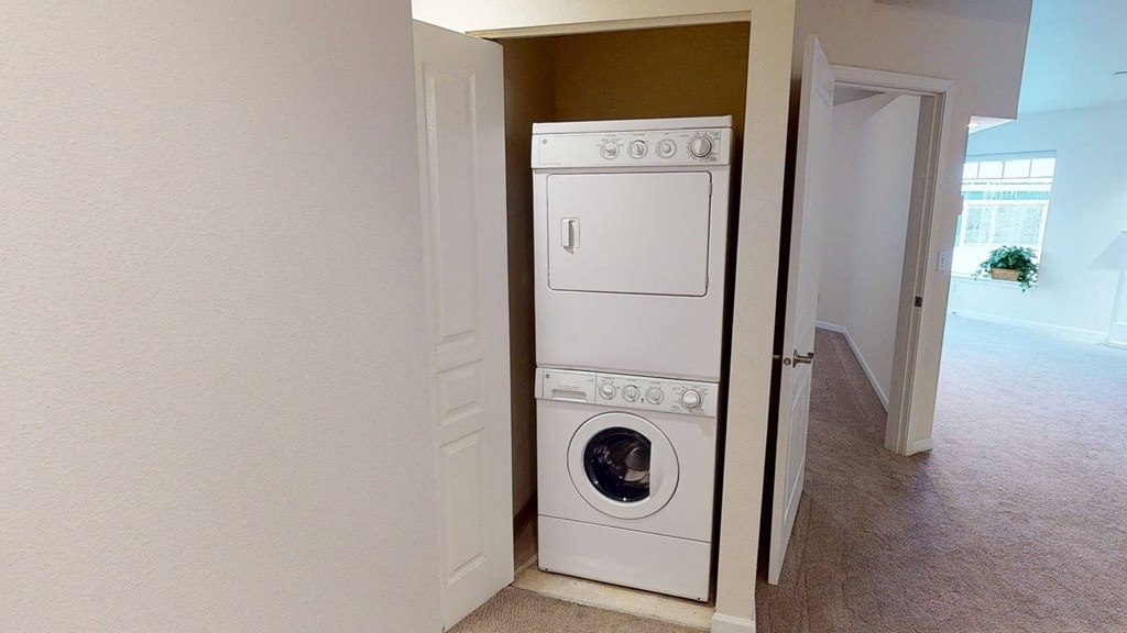 Stacked washer and dryer in a small open closet, with adjacent carpeted hallway and bright room with a large window and potted plant in the background. Located at Admirals Cove Apartments.
