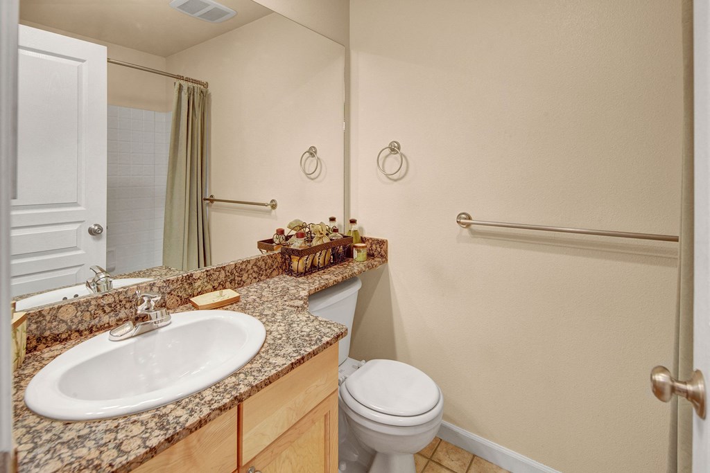 Compact bathroom with beige walls, white sink, and toilet. Granite countertop holds decorative items. Neutral shower curtain and metal towel bars. Clean and tidy ambiance. Located at Admirals Cove Apartments.