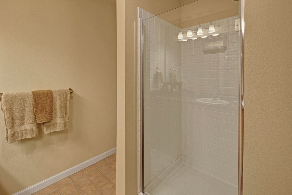 Modern bathroom with beige tiles and walls, featuring a glass shower with white tiling. Beige and brown towels hang on a towel rack, creating a cozy feel. Located at Admirals Cove Apartments.