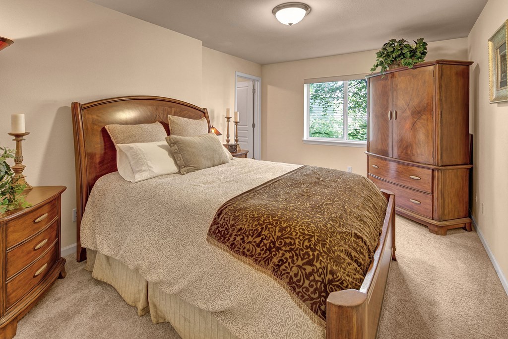 Admirals Cove Apartments - Bedroom Cozy bedroom with a large wooden bed, ornate pillows, and a patterned throw blanket. Wooden dresser and plants by a window create a warm, inviting ambiance. Located at Admirals Cove Apartments.
