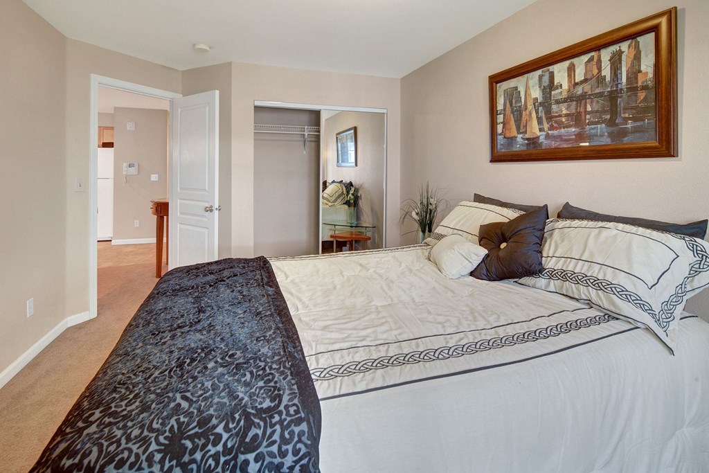 Cozy Admirals Cove Apartment bedroom with a neatly made bed featuring patterned sheets and pillows. A large painting hangs above the bed, and a closet with mirrored doors is open. Soft neutral tones create a calm ambiance.