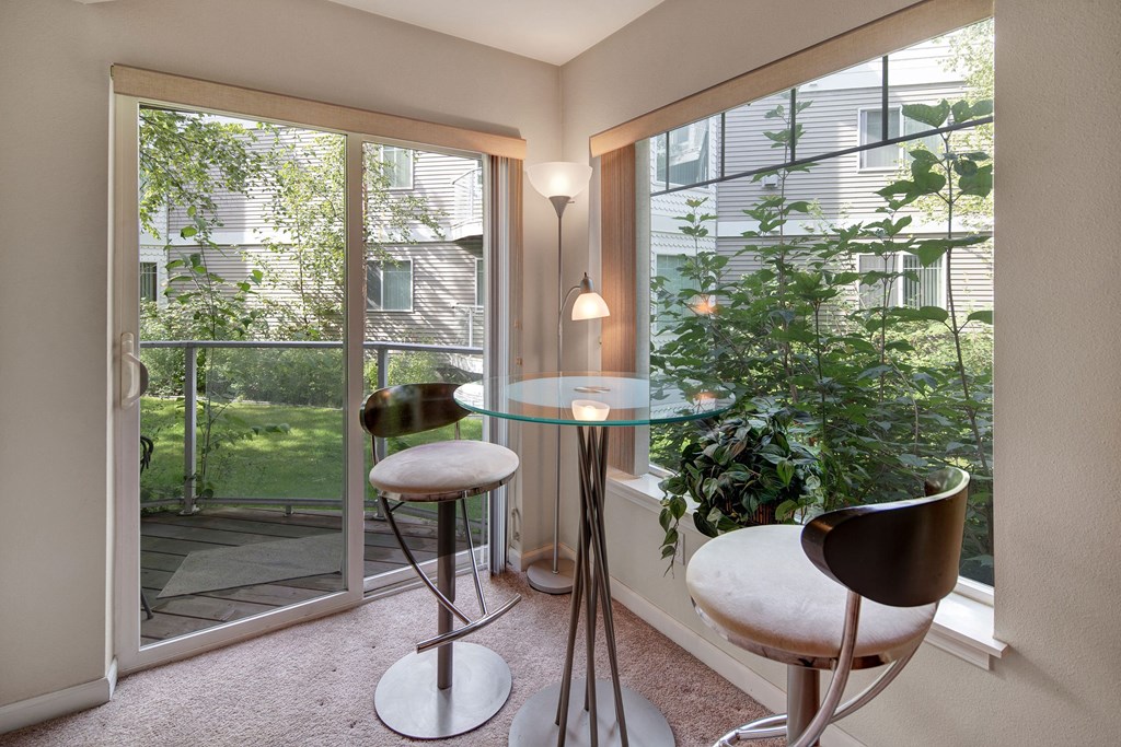 Admirals Cove Apartments - Dining Area Modern dining nook with a glass table and two sleek metal stools, beside large windows overlooking lush greenery. Bright, serene atmosphere. Located at Admirals Cove Apartments.