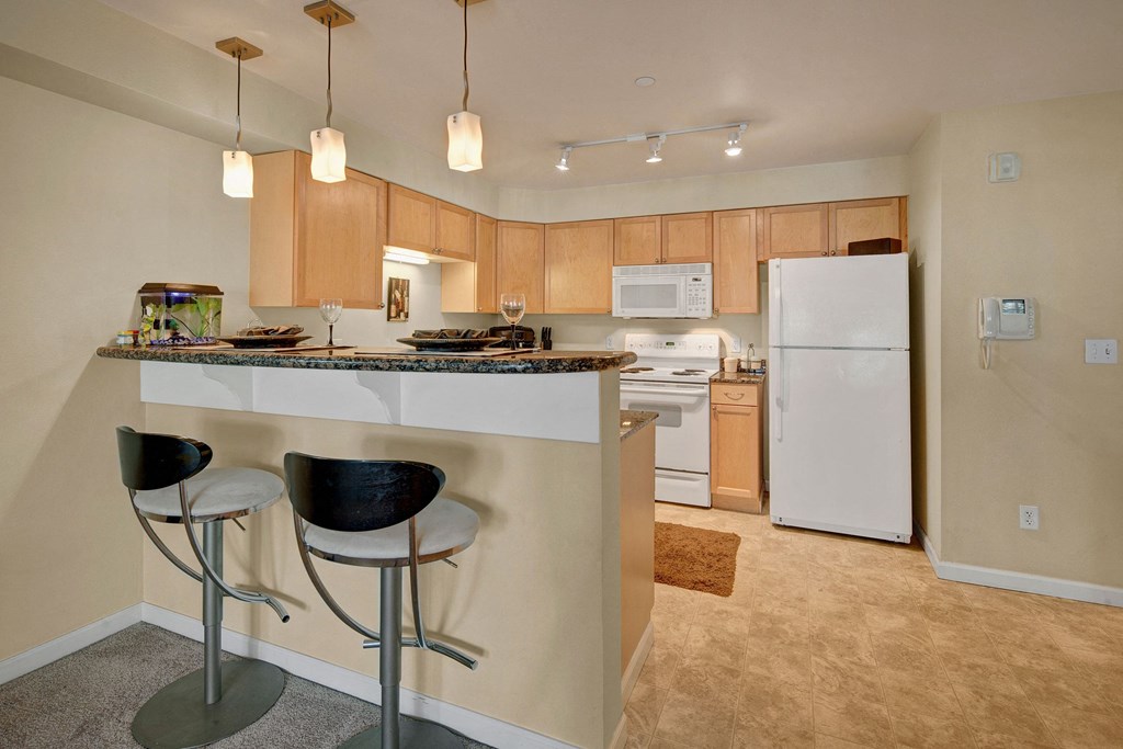 Compact kitchen with light wood cabinets, white appliances, and a small dining bar with two stools. Hanging lights illuminate the space, and a fishbowl sits on the counter at Admirals Cove Apartments