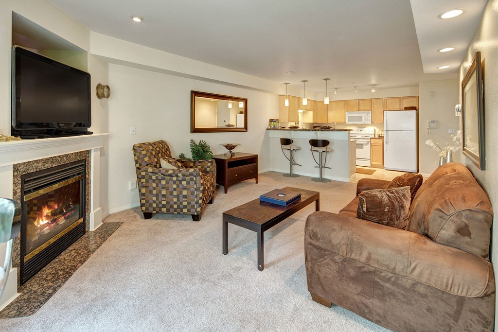 Cozy living room at Admirals Cove with a lit fireplace, wall-mounted TV, and patterned armchair. Open kitchen with bar stools and modern appliances. Warm tones.
