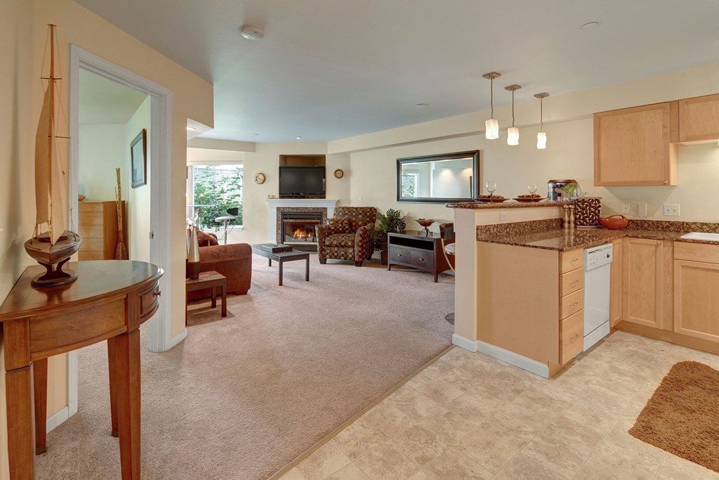 Spacious Admirals Cove living room with beige walls and carpet, featuring a cozy fireplace, brown sofa, patterned armchair, TV, and an adjacent kitchen with a granite countertop
