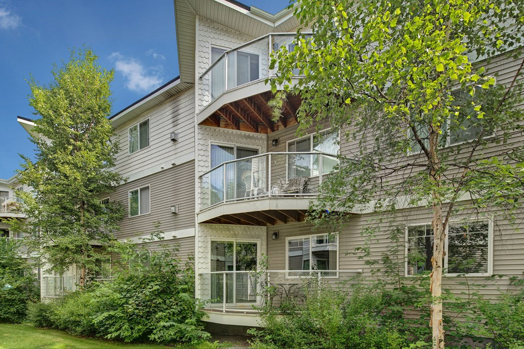 Admirals Cove three-story apartment building with balconies, surrounded by lush green trees and bushes. Bright, sunny day with a clear blue sky, conveying tranquility.