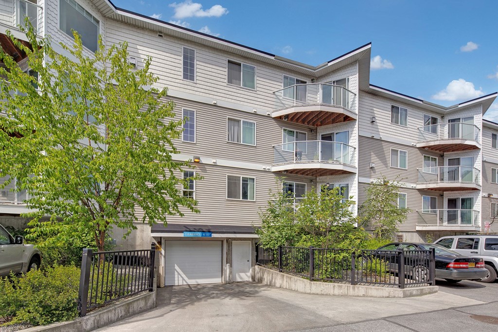 Admirals Cove four-story apartment building with white siding and glass balconies under a clear sky. Green trees and parked cars are in the foreground.