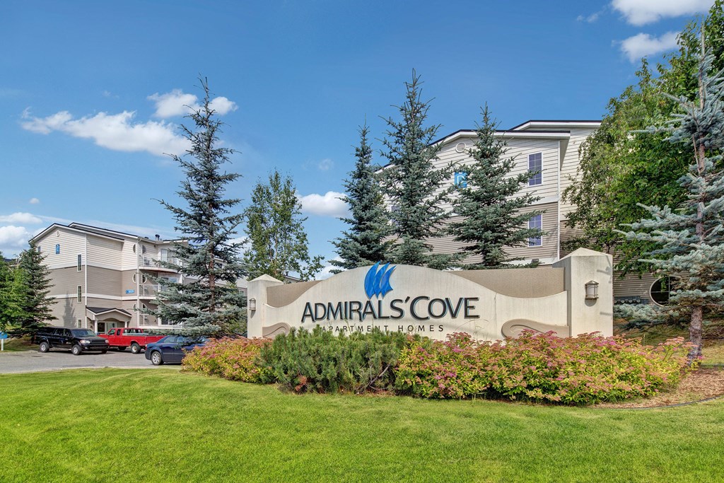 Sign for Admirals Cove Apartment Homes is surrounded by lush greenery and pine trees. Modern buildings are visible in the background under a clear blue sky.