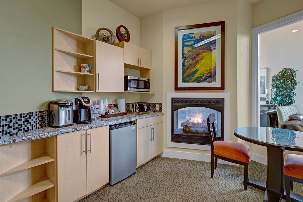 Alpine Apartments - Lounge Cozy kitchen in the community lounge with light wood cabinets, granite countertop, microwave, and coffee maker. A small fridge, glass table, and lit fireplace add warmth. Located at The Alpine Apartments.