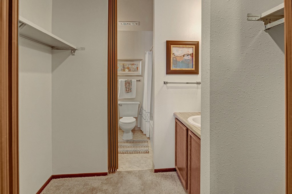A narrow bathroom with beige walls features a toilet and sink. Artwork and a towel hang on the walls, while a striped rug adds subtle color. Located at Alpine Apartments.