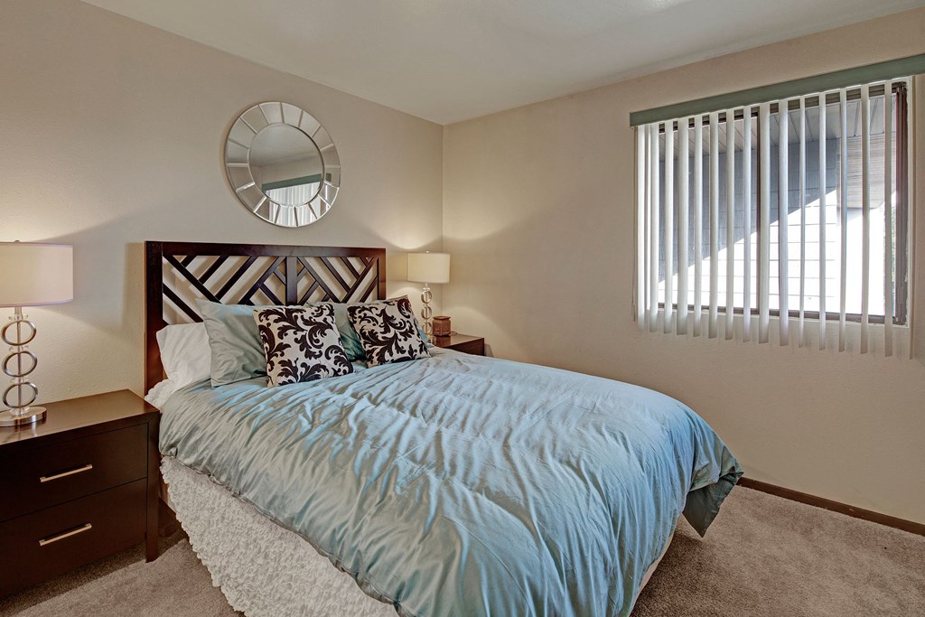 Alpine Apartments - Bedroom Alpine Apartments. Cozy bedroom with a large bed featuring a blue duvet and patterned pillows. Two bedside tables have lamps. A circular mirror hangs on the wall.