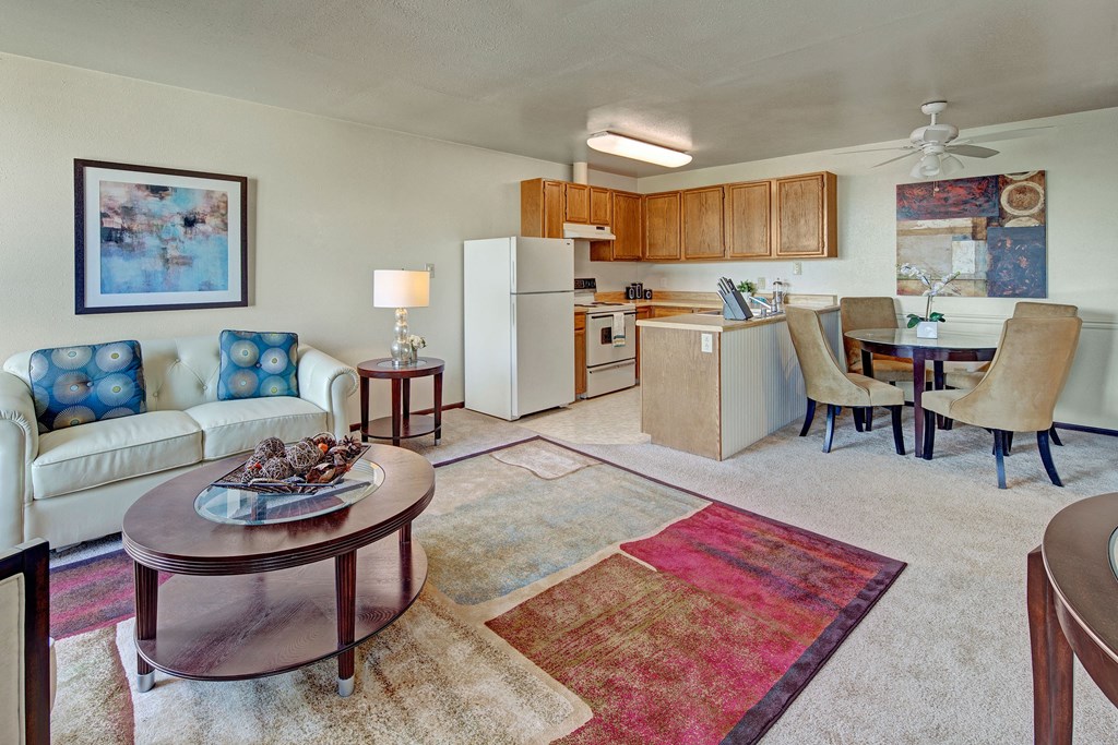 A cozy living and kitchen area with beige walls and carpet. A white sofa hosts patterned cushions beside a round table with a lamp. The kitchen features wood cabinets, and a dining area with a round table and tan chairs. A colorful abstract rug and artwork add warmth. Located at Alpine Apartments.