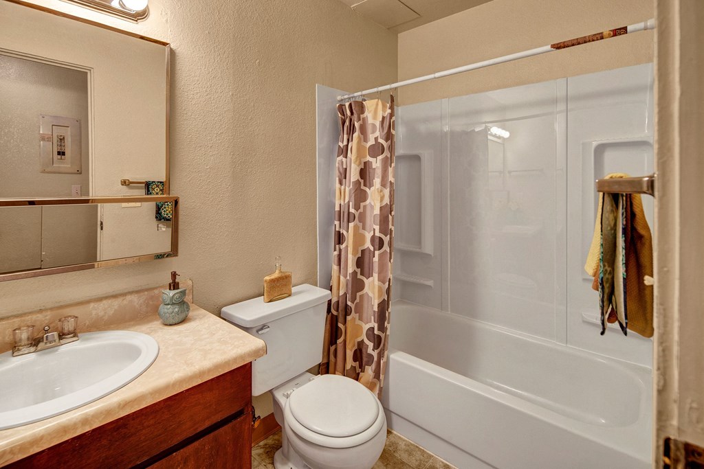 A small bathroom with beige walls, a countertop sink, toilet, and a bathtub with a patterned shower curtain. A mirror and towel add a cozy touch. Arctic Sun Apartments.
