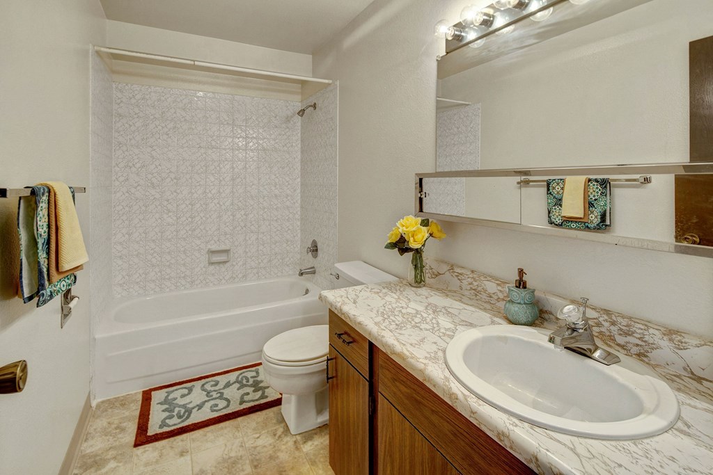 Bright bathroom with white tiled bathtub, brown marble sink, and large mirror. Yellow roses in vase, towels, and decorative soap add color. Cozy and neat. Arctic Sun Apartments.